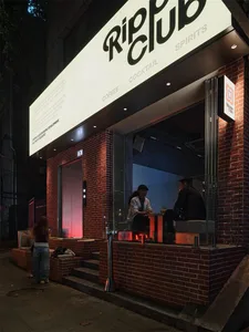 Night view of Ripple Club bar with red brick exterior, large illuminated sign, and two patrons seated at a warmly lit counter inside.