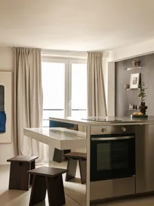 Modern minimalist kitchen featuring a white tiled breakfast bar, built-in oven, walnut wood stools, and large windows with linen curtains allowing abundant natural light.