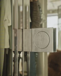 Close-up of a brushed stainless steel closet rod system featuring a square geometric bracket with a circular detail, with hanging clothes visible in the background.