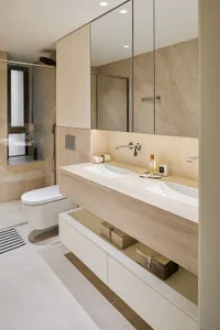 Modern minimalist bathroom with a long floating travertine double vanity, two white ceramic sinks, chrome wall-mounted faucets, large frameless mirror cabinet, and a white wall-hung toilet visible in the background.