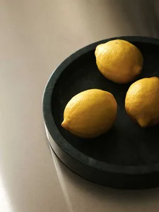 Close-up of three whole yellow lemons placed on a round dark green marble tray against a neutral textured background.