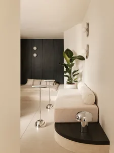 Modern minimalist living room with a built-in boucle sofa, dark wood accent wall, chrome side tables, and a large potted plant in a bright, neutral space.