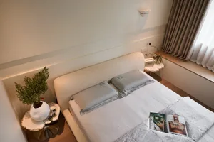 High-angle view of a modern minimalist bedroom with a cream boucle upholstered bed, grey silk pillows, marble side tables, and large window with neutral curtains.