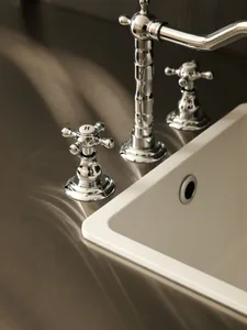 Close-up of a polished chrome bathroom faucet with white ceramic cross handles marked with H and C, mounted on a smooth stone countertop next to a white ceramic sink.