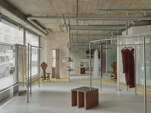 Modern minimalist boutique interior with raw concrete walls, large windows, steel clothing racks displaying red and white dresses, sculptural wooden chairs, and a curved yellow floor area under an exposed ceiling with visible ductwork.