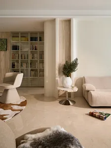 Modern living room featuring a travertine-topped pedestal side table with a potted plant, a beige swivel armchair on a cowhide rug, built-in floor-to-ceiling bookshelves, and a curved beige sofa under warm natural light.
