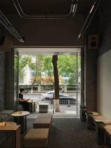 Modern minimalist cafe interior with concrete walls, wooden benches, beige ottomans, round tables, and a large open facade framing a street tree and passing cars.