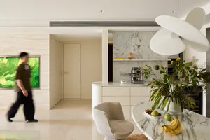 Modern open-plan kitchen and living area featuring a curved Calacatta marble island, sculptural white pendant light, built-in coffee station against a marble backsplash, and a beige boucle swivel chair.