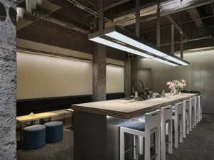Modern industrial-style commercial bar interior with long concrete counter, white bar stools, blue ottomans, and exposed ceiling infrastructure.