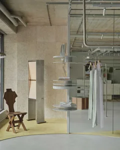Modern boutique interior featuring raw concrete walls, a circular yellow rug, a vintage wooden folding chair, and a multi-tiered metal display stand with books and hanging garments.