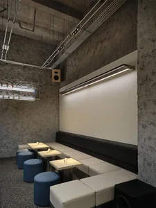 Modern restaurant interior featuring raw concrete walls, built-in white upholstered banquette, small square tables, blue cylindrical ottomans, and dramatic linear LED lighting.