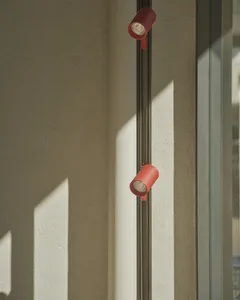 Two red cylindrical track spotlights mounted vertically on a black track against a textured concrete wall with strong diagonal sunlight creating sharp shadows.