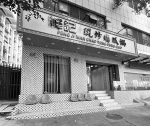 Black and white photograph of a retro-style restaurant storefront with black and white mosaic tile walls, four plastic stools on the steps, vertical metal security gate, and large illuminated sign reading Feng Ji Xian Chao Tong Feng Guo.
