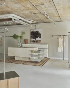 Modern minimalist boutique interior featuring a large travertine stone central island, white modular cabinetry, industrial track lighting, and clothing racks under an exposed concrete ceiling.