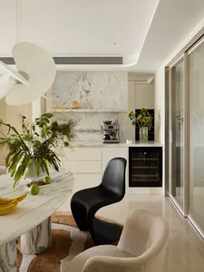 Modern open kitchen and dining area with Calacatta marble round table, black organic-shaped chair, beige bouclé armchair, and marble backsplash with built-in coffee station.