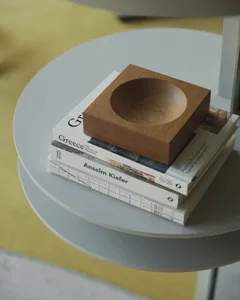 Close-up of a solid oak decorative bowl placed on a stack of books including titles about Greece and Anselm Kiefer, resting on a light grey round side table with a yellow rug in the background.