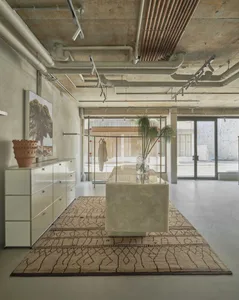 Modern minimalist boutique interior featuring a large onyx marble central island on a patterned rug, white modular cabinetry, concrete walls and ceiling with exposed white and copper pipes, and large glass doors at the back.