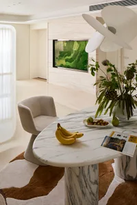 Modern dining area featuring a white and grey Calacatta marble oval table, a beige boucle swivel chair, a large abstract green artwork on a travertine wall, and a brown and white abstract rug.