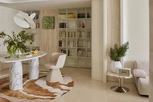 Modern minimalist living and reading area featuring a white marble round table on a brown and white cowhide rug, a sculptural white pendant lamp, built-in light wood bookshelves, and travertine accents.