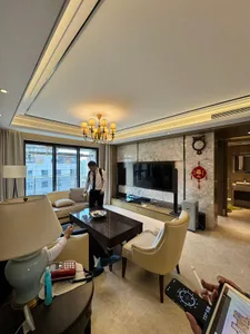 Modern luxury living room with beige leather sofas, dark wood coffee table, marble TV wall, crystal chandelier, and large windows overlooking city buildings.