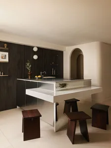 Modern minimalist kitchen featuring a walnut and stainless steel island with white tiled overhang and dark wooden block stools in a bright, neutral space with an arched doorway.