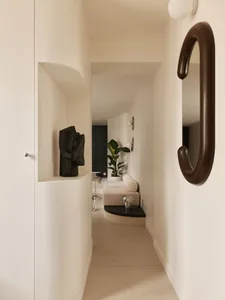 Narrow modern hallway featuring a curved built-in boucle window seat, potted plant, sculptural black artwork in a wall niche, and an organic-shaped brown mirror.