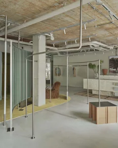 Modern boutique interior with curved stainless steel clothing display racks, polished concrete floor, exposed ceiling infrastructure, a shaggy brown armchair on a yellow rug, and a marble checkout counter.