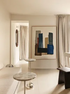 Modern minimalist living room featuring a large abstract watercolor painting in blue, black, and earth tones, two round marble side tables with chrome bases, a cream bouclé sofa, and beige linen curtains.