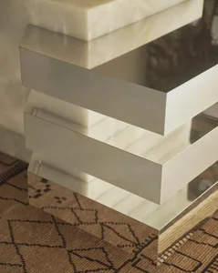 Close-up of a modern stepped side table made of creamy onyx marble with polished stainless steel edges, placed on a brown geometric-patterned rug.