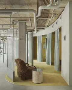 Modern fitting room area with curved concrete wall, exposed ceiling pipes, chartreuse carpet, brown sheepskin-covered armchair and marble side table