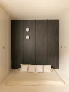 Minimalist Japanese-style bedroom featuring a floor-level tatami bed with beige linen bedding, a large dark wood paneled wall, and two spherical white wall sconces.