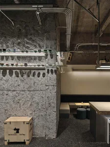 Industrial-style café interior featuring a textured raw concrete wall, two tiers of floating glass shelves displaying ceramic tableware, exposed ceiling ducts, and minimalist seating area with wooden counter and upholstered benches.