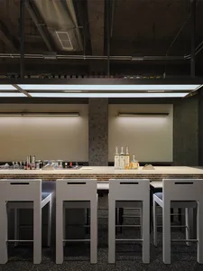 Modern industrial bar counter with four white minimalist stools, concrete walls, and overhead linear LED lighting in a commercial space.
