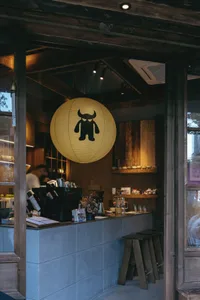 View into a cozy café with a large yellow paper lantern featuring a black monster graphic hanging above a concrete and wood coffee bar counter, wooden stools, and warm track lighting.