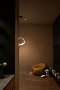 Modern minimalist reading nook with a mustard yellow curved lounge chair, irregular glass coffee table, stacked books, and a glowing circular pendant light in a warm earth-toned space.