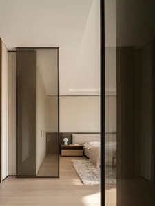 View into a minimalist modern bedroom with light oak wood flooring, neutral beige walls, a textured fabric headboard, floating nightstand, and abstract area rug.
