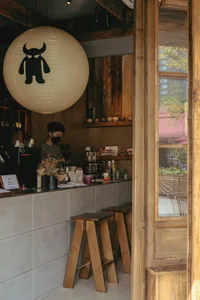 View of a minimalist cafe counter with concrete block base, wooden stools, a barista working, and a large white paper lantern with a black monster graphic hanging from the ceiling.