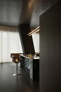 Modern minimalist kitchen featuring a large dark marble island, a sculptural bronze bar stool, dramatic angular architecture, and sheer curtains in the background.