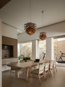 Modern minimalist dining room featuring a long solid oak table, eight matching oak dining chairs with light upholstery, two large wooden artichoke pendant lights, white walls, and a dramatic natural stone feature wall visible through large openings, with a kitchen counter and treadmill in the background.