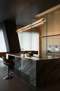 Modern luxury kitchen featuring a large dark marble island with integrated cooktop and sink, a transparent amber bar stool, and a long linear gold pendant light hanging above.