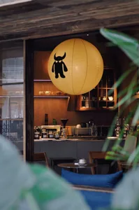 Interior view of a cozy cafe featuring a large yellow paper lantern with a black monster design hanging above a wooden coffee counter with warm lighting and wooden furniture.