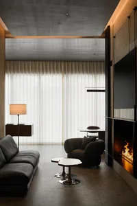 Modern minimalist living room featuring a dark grey concrete ceiling, sheer curtains, black leather sofa, boucle armchair, live-edge coffee table, and a built-in fireplace with visible flames.