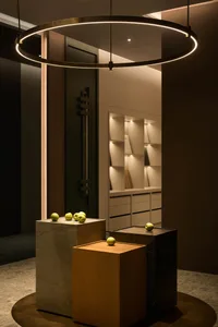 Modern minimalist entryway featuring a large circular LED pendant lamp, three display pedestals in concrete, light oak, and black marble, each topped with green apples, with built-in illuminated shelving in the background.