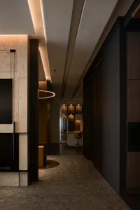 Modern luxury hallway with dark wood paneling, travertine stone wall, circular pendant light, and view into a backlit display cabinet and kitchen area.