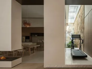 Modern open-plan interior showing a dining area, kitchen, fireplace, wooden pendant light, and a treadmill facing a stone-walled courtyard with skylight.