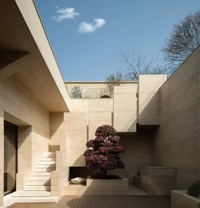Modern minimalist courtyard with beige travertine stone walls, wooden deck, concrete stairs, and a vibrant red-flowering bonsai tree under a clear blue sky.