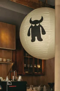Interior view of a specialty coffee bar featuring warm wooden cabinetry, open shelving with ceramic cups, and a large white paper lantern with a black monster illustration hanging prominently in the foreground.