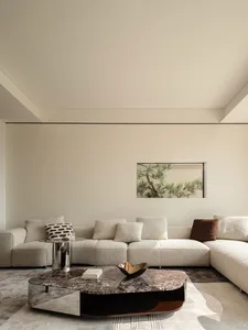 Modern minimalist living room featuring a large cream boucle L-shaped sectional sofa, a dark marble and chrome coffee table, and a framed pine tree artwork on a beige wall.