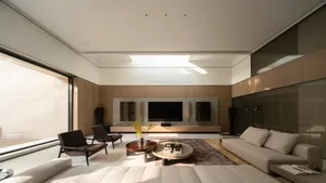 Modern minimalist living room with beige sofa, dark brown leather armchairs, round coffee tables on a rug, and a large walnut entertainment center with wall-mounted TV under a skylight.