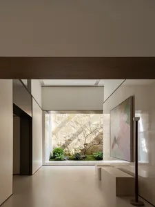 Modern minimalist interior hallway with light stone floors, wooden ceiling beams, built-in benches, and a large abstract painting, opening to a Japanese-style courtyard with moss, ferns, a small tree, and a dramatic natural stone wall.